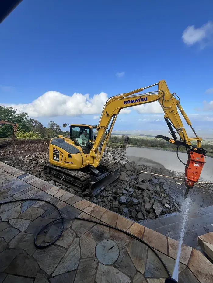 Excavator with a hydraulic breaker demolishing a swimming pool surrounded by flagstone decking