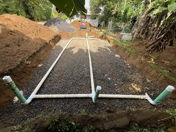 Septic disposal bed of drain rock with white distribution pipe and green cleanout risers, a mini excavator at the far end of the trench