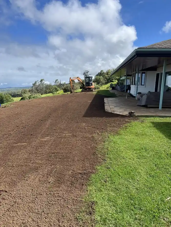 Freshly graded red-dirt slope beside a house and lawn with an excavator working uphill